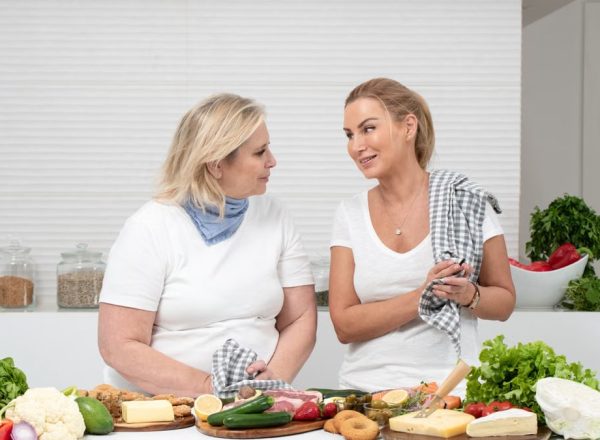 Two women in a kitchen preparing fresh vegetables and cheese with olive oil and spices.